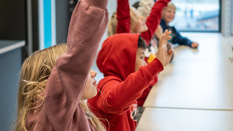 Elever sitter längs ett bord i klassrum och räcker upp händerna